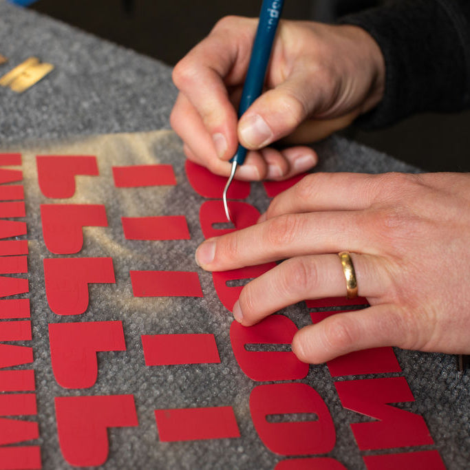 Hands weeding red vinyl letters on a grey workshop bench.