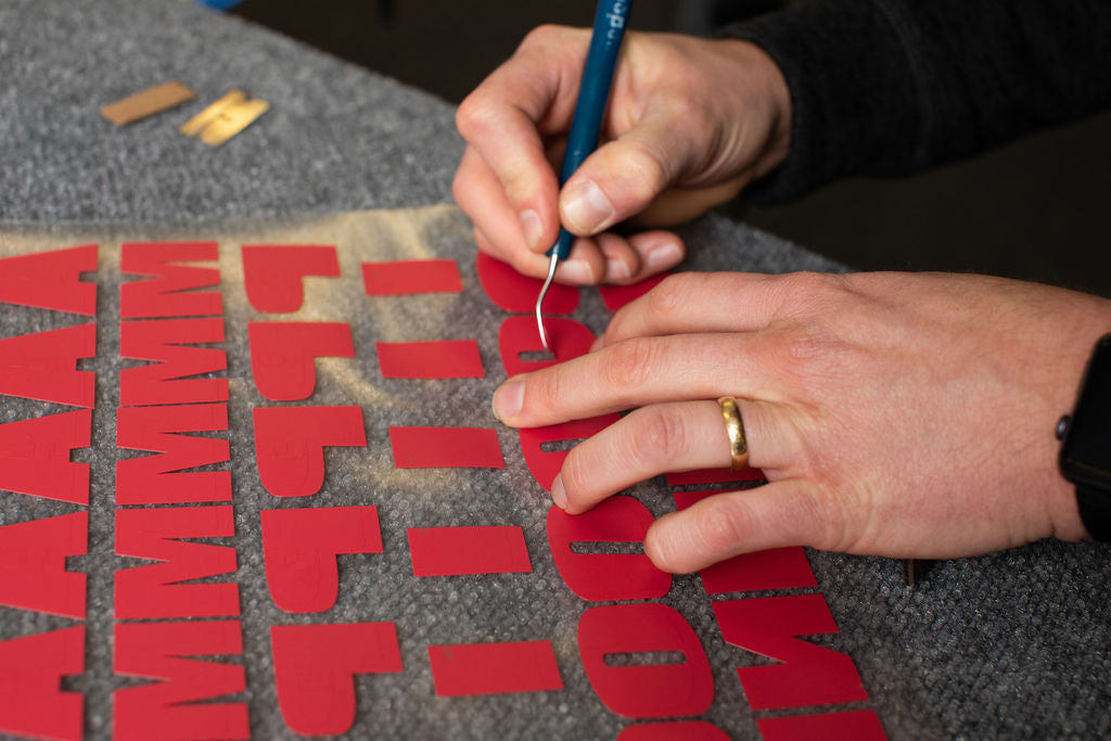 Hands weeding red vinyl letters on a grey workshop bench.
